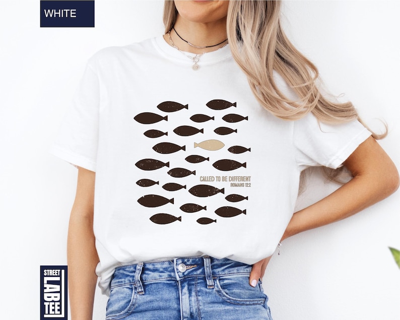 Puede incluir: Camiseta blanca con un gr&aacute;fico de peces marrones, uno beige, nadando juntos. El texto "CALLED TO BE DIFFERENT ROMANS 12:2" est&aacute; impreso debajo. La camiseta est&aacute; etiquetada como "WHITE" en la esquina superior izquierda.