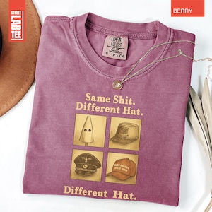 May include: Berry-colored t-shirt with the text "Same Shit. Different Hat." and four hat illustrations. A gold necklace with a pendant is on the shirt. The shirt has a Comfort Colors tag. A brown hat and a dried leaf are in the background.