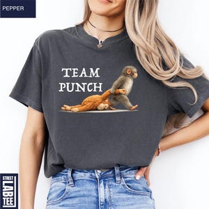 May include: Dark gray t-shirt with the text "TEAM PUNCH" in white. The shirt features a cartoon illustration of two monkeys, one standing and the other lying down. The shirt is a comfortable, casual style.