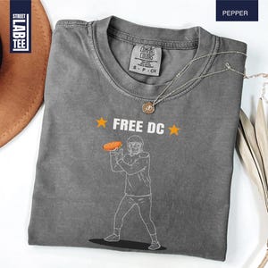 Op de afbeelding: Een grijs t-shirt met de tekst "FREE DC" en een afbeelding van een American footballspeler die een hotdog gooit. Het shirt heeft een ronde hals en is gemaakt van zacht materiaal. Een gouden ketting is over het shirt gedrapeerd.