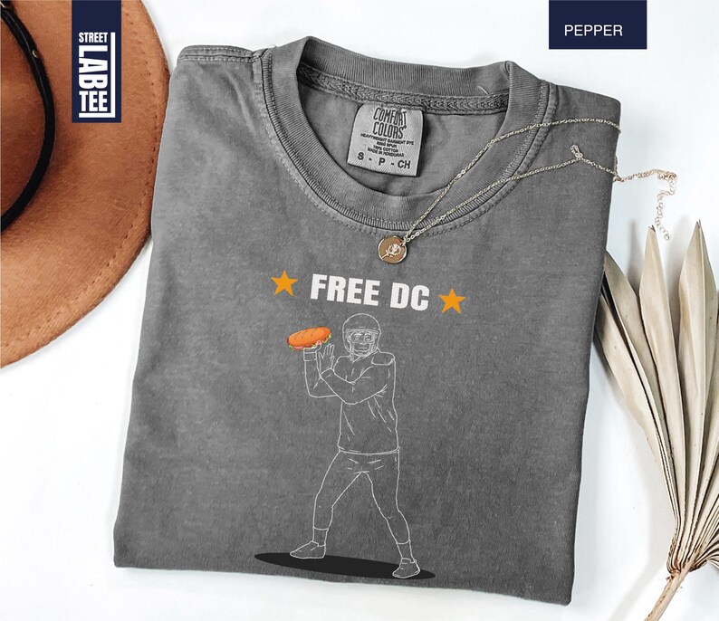 Op de afbeelding: Een grijs t-shirt met de tekst "FREE DC" en een afbeelding van een American footballspeler die een hotdog gooit. Het shirt heeft een ronde hals en is gemaakt van zacht materiaal. Een gouden ketting is over het shirt gedrapeerd.