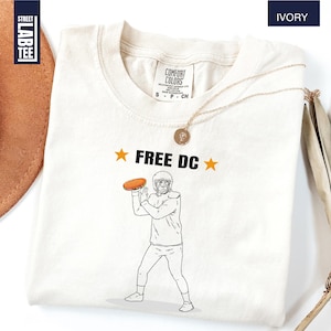 Op de afbeelding: Ivoor kleurig t-shirt met de tekst "FREE DC" en een afbeelding van een American footballspeler die een bal gooit. Het shirt heeft een ronde hals en korte mouwen. Het shirt is gemaakt van een zacht materiaal.