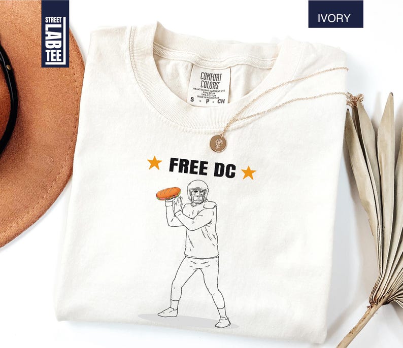 Op de afbeelding: Ivoor kleurig t-shirt met de tekst "FREE DC" en een afbeelding van een American footballspeler die een bal gooit. Het shirt heeft een ronde hals en korte mouwen. Het shirt is gemaakt van een zacht materiaal.