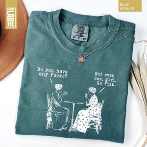 May include: Blue Spruce t-shirt with the text "Do you have any fucks?" and "Not even one, girl. Go fish." in white. The shirt features a graphic of two women at a table. A gold necklace is draped on the shirt.
