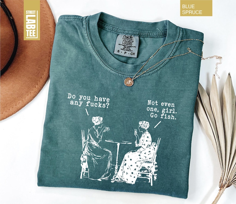 May include: Blue Spruce t-shirt with the text "Do you have any fucks?" and "Not even one, girl. Go fish." in white. The shirt features a graphic of two women at a table. A gold necklace is draped on the shirt.