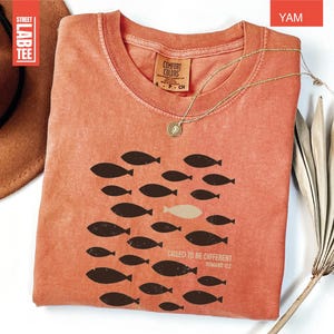 Puede incluir: Una camiseta color &ntilde;ame con cuello redondo y mangas cortas. La camiseta presenta un gr&aacute;fico de peces, con un pez de un color diferente. El texto "CALLED TO BE DIFFERENT ROMANS 12:2" est&aacute; impreso en la camiseta. Un collar de oro est&aacute; sobre la camiseta.