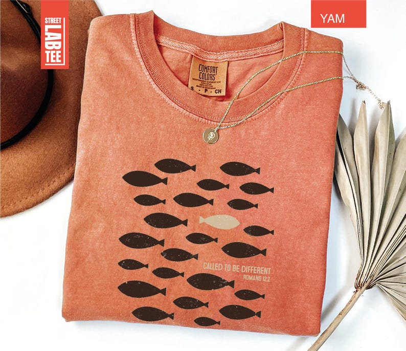 Puede incluir: Una camiseta color &ntilde;ame con cuello redondo y mangas cortas. La camiseta presenta un gr&aacute;fico de peces, con un pez de un color diferente. El texto "CALLED TO BE DIFFERENT ROMANS 12:2" est&aacute; impreso en la camiseta. Un collar de oro est&aacute; sobre la camiseta.