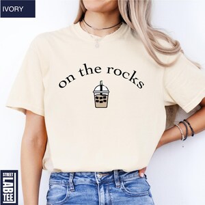 May include: Ivory-coloured t-shirt featuring the text "on the rocks" above an iced drink cartoon. The shirt has a crew neck and is a neutral colour. The shirt is made of a soft material.