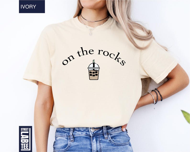 May include: Ivory-coloured t-shirt featuring the text "on the rocks" above an iced drink cartoon. The shirt has a crew neck and is a neutral colour. The shirt is made of a soft material.