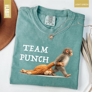 May include: Light green t-shirt with the words "TEAM PUNCH" in white block letters. The shirt features a graphic of a monkey pulling another monkey. A silver necklace with a pendant is on the shirt.
