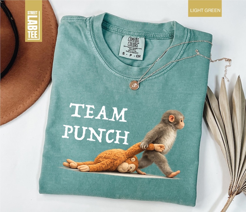 May include: Light green t-shirt with the words "TEAM PUNCH" in white block letters. The shirt features a graphic of a monkey pulling another monkey. A silver necklace with a pendant is on the shirt.