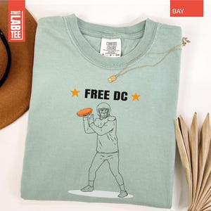 Op de afbeelding: Lichtgroen t-shirt met de tekst "FREE DC" en een afbeelding van een American footballspeler die een bal gooit. Het shirt heeft een ronde hals en korte mouwen. Een gouden ketting ligt over het shirt.