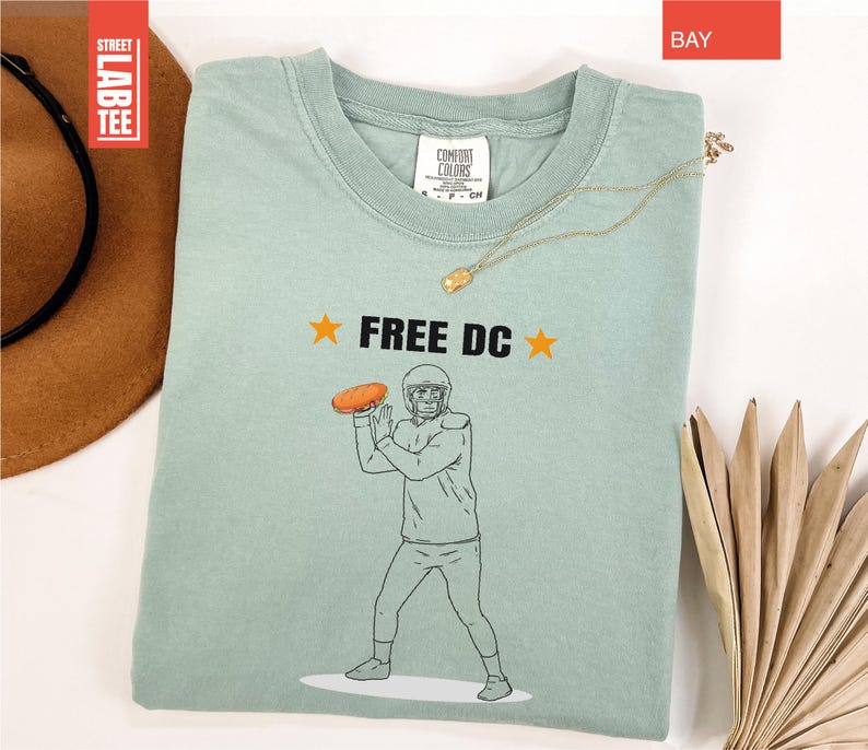 Op de afbeelding: Lichtgroen t-shirt met de tekst "FREE DC" en een afbeelding van een American footballspeler die een bal gooit. Het shirt heeft een ronde hals en korte mouwen. Een gouden ketting ligt over het shirt.