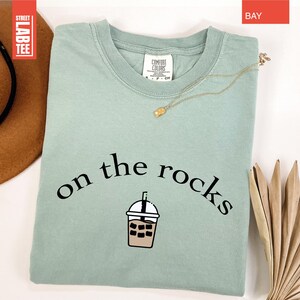 May include: A pale green t-shirt featuring the words "on the rocks" and an iced coffee graphic. A gold necklace is draped across the folded shirt. The shirt has a tag that says "Comfort Colors".