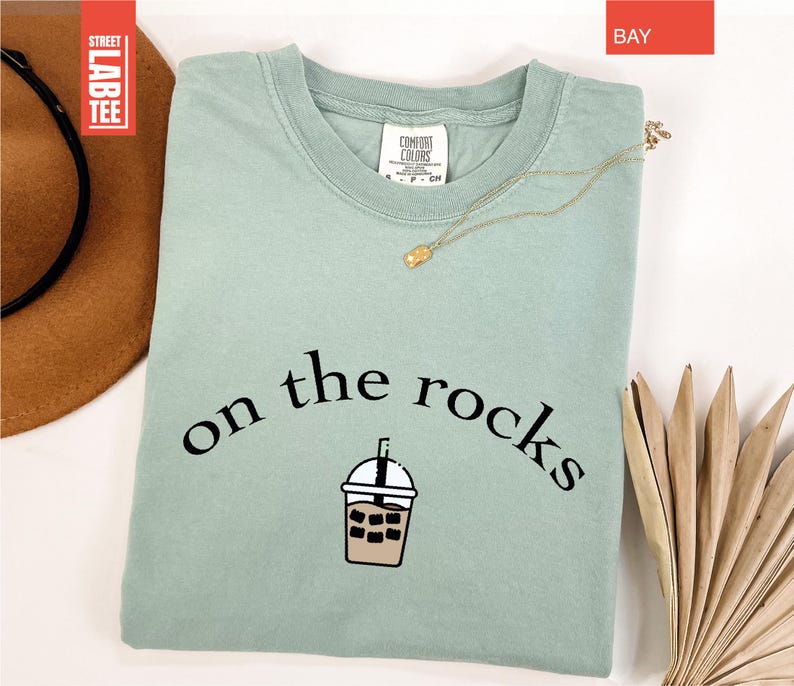 May include: A pale green t-shirt featuring the words "on the rocks" and an iced coffee graphic. A gold necklace is draped across the folded shirt. The shirt has a tag that says "Comfort Colors".