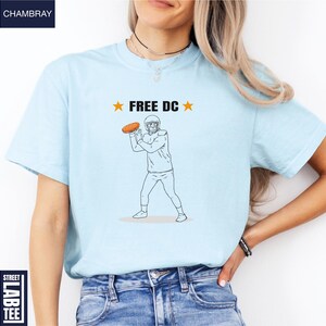 Op de afbeelding: Lichtblauw T-shirt met een afbeelding van een footballspeler die een bal gooit, en de tekst "FREE DC" met sterren. Het shirt heeft een ronde hals en is gemaakt van zacht materiaal.