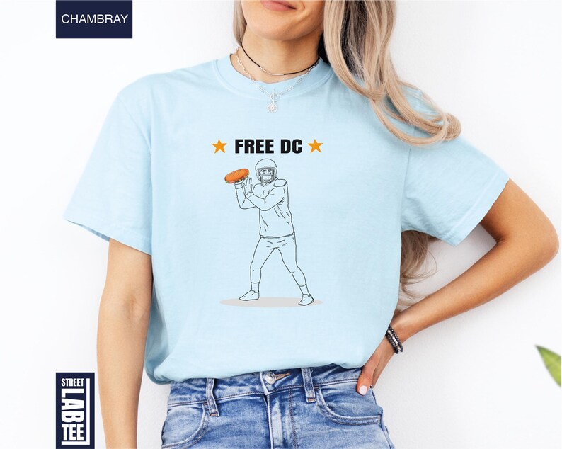 Op de afbeelding: Lichtblauw T-shirt met een afbeelding van een footballspeler die een bal gooit, en de tekst "FREE DC" met sterren. Het shirt heeft een ronde hals en is gemaakt van zacht materiaal.
