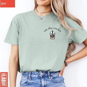 May include: A light green t-shirt with the words "on the rocks" above a graphic of an iced coffee. The shirt is a solid color and has short sleeves. The shirt is paired with blue jeans.