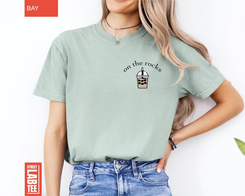 May include: A light green t-shirt with the words "on the rocks" above a graphic of an iced coffee. The shirt is a solid color and has short sleeves. The shirt is paired with blue jeans.