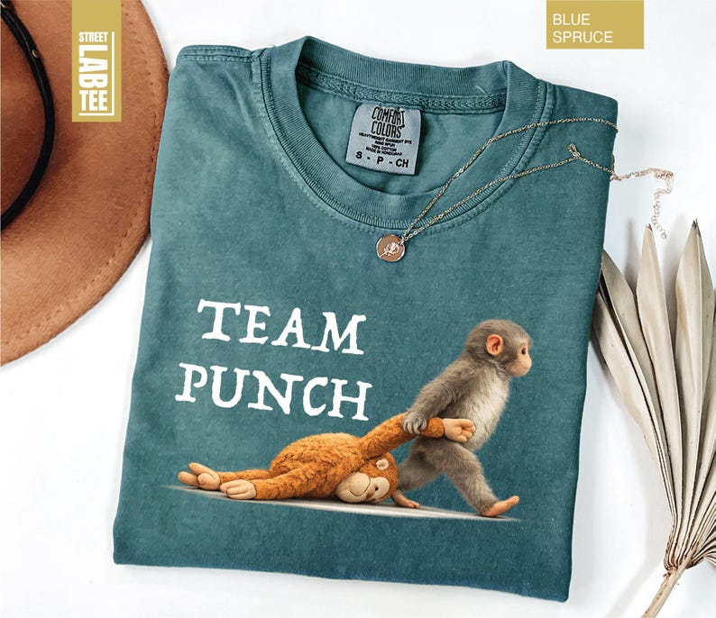 May include: Blue spruce Comfort Colors t-shirt with the text "TEAM PUNCH" and a graphic of a monkey pulling a stuffed animal. The shirt has a crew neck and short sleeves. A gold necklace with a pendant is on the shirt.