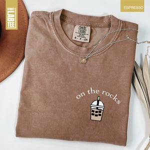 May include: Espresso-colored t-shirt with the phrase "on the rocks" and a graphic of a bubble tea drink. The shirt has a crew neck and short sleeves. A silver necklace with a small pendant is draped across the shirt.