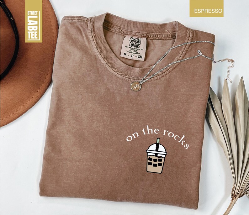 May include: Espresso-colored t-shirt with the phrase "on the rocks" and a graphic of a bubble tea drink. The shirt has a crew neck and short sleeves. A silver necklace with a small pendant is draped across the shirt.