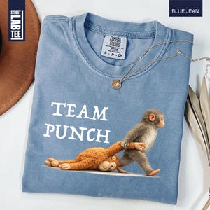 May include: Blue jean colored t-shirt with the words "TEAM PUNCH" in white. The shirt features a graphic of two monkeys, one pulling the other. A gold necklace and a brown hat are also in the image.