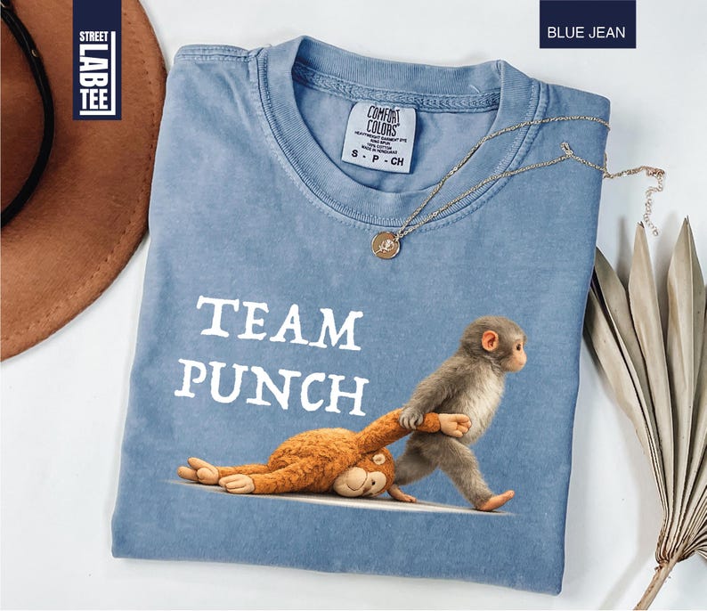 May include: Blue jean colored t-shirt with the words "TEAM PUNCH" in white. The shirt features a graphic of two monkeys, one pulling the other. A gold necklace and a brown hat are also in the image.