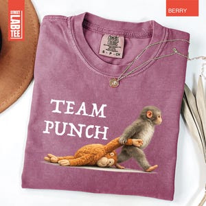 May include: Berry-colored t-shirt with the text "TEAM PUNCH" and a graphic of two monkeys. One monkey is standing and pulling the other. A gold necklace is draped on the shirt. The shirt has a crew neck and short sleeves.