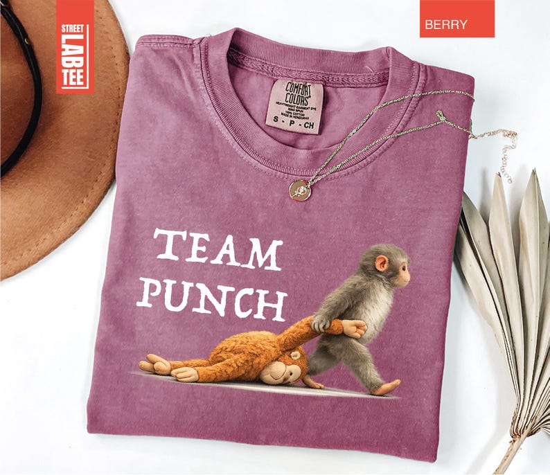 May include: Berry-colored t-shirt with the text "TEAM PUNCH" and a graphic of two monkeys. One monkey is standing and pulling the other. A gold necklace is draped on the shirt. The shirt has a crew neck and short sleeves.