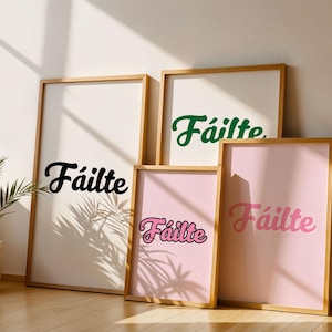 May include: Four framed art prints displaying the word "Fáilte" in black, green, pink, and a pink and black gradient. The prints are in wooden frames and lean against a white wall. A potted plant is visible in the corner.