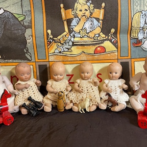 May include: Six vintage porcelain dolls with painted features, four wearing cream-colored crocheted dresses, positioned alongside two red wooden toy trains with green polka dots, a small wooden figurine, a plastic animal, and black binoculars.