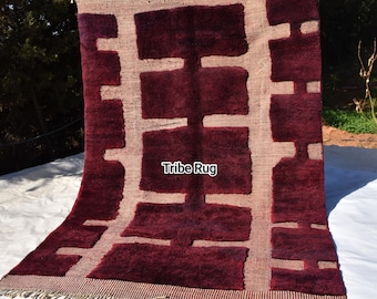 May include: A rectangular rug featuring a deep burgundy, high-pile geometric pattern set against a light beige, textured woven background. The rug includes the text Tribe Rug in the center and has a fringed edge along the bottom border.
