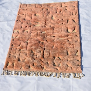 May include: A peach-coloured rectangular rug with a textured, geometric pattern. The rug has a soft, plush appearance and is trimmed with a fringe along the bottom edge. The rug's design features raised, rounded shapes.