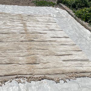 May include: A cream-coloured rug with a textured, wavy pattern and fringe along the edges. The rug is laid out on a white plastic sheet.