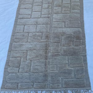 May include: A rectangular beige rug with a brick-like pattern. The rug has a fringe along the bottom edge. The design features various rectangular shapes, creating a textured appearance. The rug is suitable for a living room or bedroom.