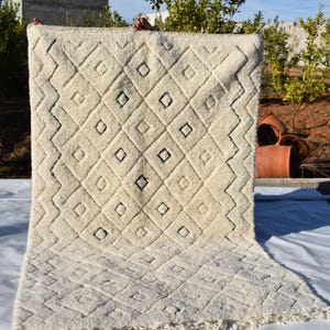 May include: A white and black Moroccan rug with a geometric diamond pattern. The rug has a fringe border and is made of wool.
