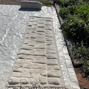 May include: A white, fluffy rug with a geometric pattern of raised squares. The rug has a fringe border and is laid out on a white surface.