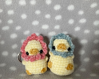 May include: Two crocheted chick keychains. One chick has a pink head covering, the other has a blue head covering. Both have yellow bodies, black eyes, and small orange feet. The keychain on the pink chick is silver.