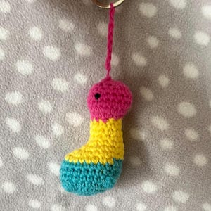 Pode incluir: Um chaveiro de crochê feito à mão em forma de figura estilizada. O chaveiro apresenta uma cabeça rosa com um pequeno olho preto, uma seção média amarela e uma base azul. Um laço de fio rosa conecta a figura a um chaveiro prateado.