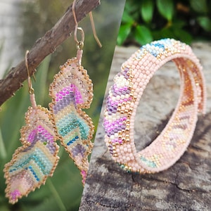 May include: Beaded feather earrings and a matching bracelet. The earrings are in a feather shape with gold-colored hooks. The bracelet and earrings feature a pastel color palette of pink, purple, blue, and gold beads. The jewelry is displayed on a wooden surface.