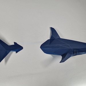 Puede incluir: Dos tiburones de origami azules, uno grande y otro pequeño, se muestran sobre un fondo blanco. Los tiburones están hechos de papel y tienen un diseño geométrico.