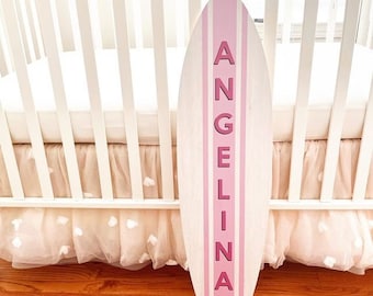 May include: A wooden surfboard-shaped sign with the name "ANGELINA" in pink lettering. The sign has pink stripes and is leaning against a white crib with a pink skirt.