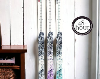 May include: Three wooden ski-shaped growth charts with a mountain scene design. The charts are painted in shades of purple, teal, and white. Each chart has a ruler marked in inches from 2 to 6 feet.