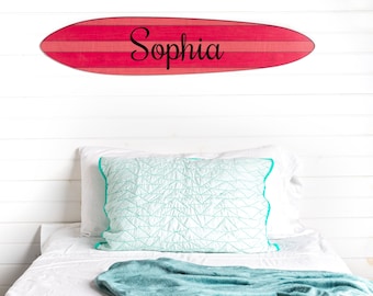 May include: A red and white surfboard with the name "Sophia" written in black lettering. The surfboard is hanging on a white wall above a white bed with a teal blanket and a white pillow with a teal geometric pattern.