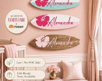 May include: Three surfboard-shaped wall plaques featuring the name Alexandra in cursive script and a hibiscus flower graphic. The top plaque is white and teal, the middle is white and pink, and the bottom is a natural wood grain with a light pink flower.