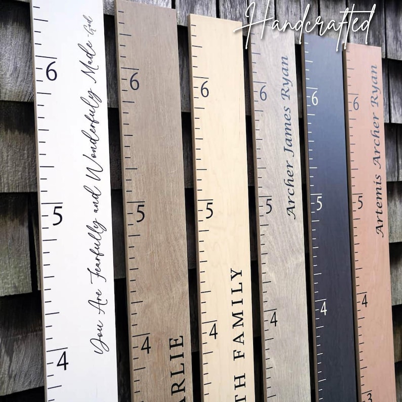Pu&ograve; includere: Sei metri da parete verticali in legno con varie finiture tra cui bianco, marrone chiaro, legno naturale, grigio, grigio scuro e rosa chiaro. Ogni tavola presenta segni di misurazione neri stampati e testo personalizzato.