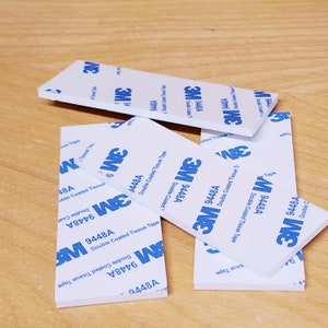 May include: A close-up of several rolls of 3M 9448A double-coated tissue tape. The tape is white with blue lettering and is designed for general purpose use.
