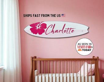 May include: A white surfboard-shaped wall decoration with a pink hibiscus flower and the name "Charlotte" in cursive. The wall art is displayed on a pink wall, with a crib visible below. Text on the wall says "Ships Fast From The US".