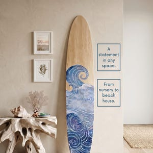 May include: A tall, narrow wooden surfboard wall decoration featuring a painted blue and white wave pattern at the bottom. The text on the right reads: A statement in any space. From nursery to beach house.